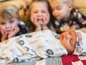 Closeup on a bed of three small children laying on their bellies and overlooking their sleeping newborn sibling, delivered by midwife Angie, in the foreground, representing midwifery care.