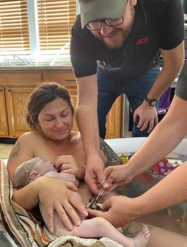 Mom holds her newborn baby in birthing tub while midwife assists dad in cutting the umbilical cord, representing birth services.